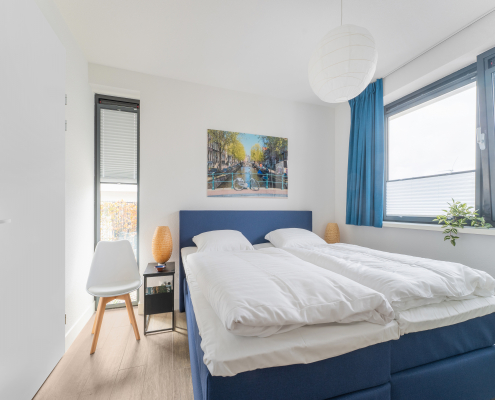 schlafzimmer mit blauem bett, konsolen mit beiger lampe, 1 stuhl, bild von Amsterdam über dem bett, fenster mit blauen gardinen